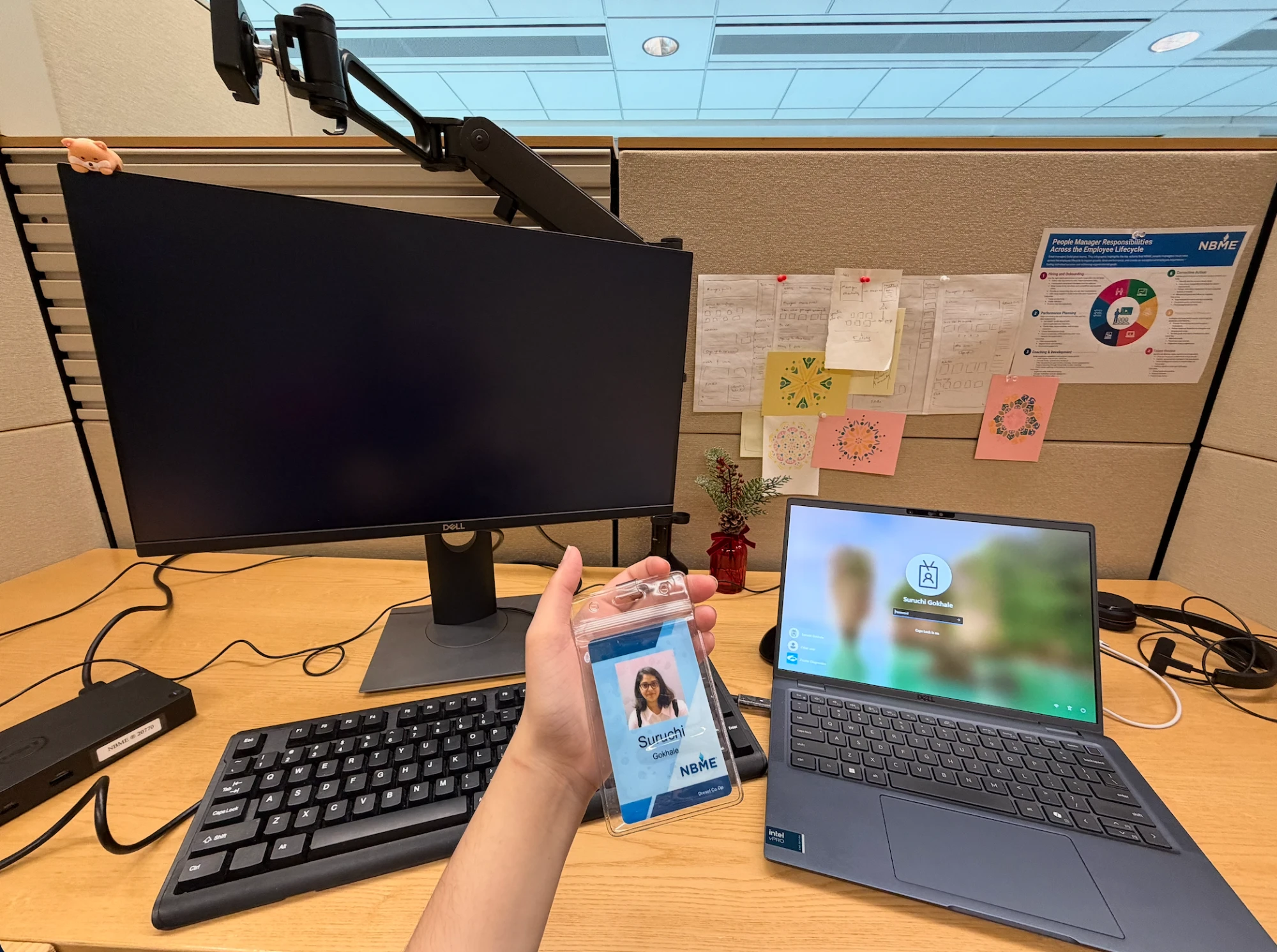 Suruchi's desk on her first day, holding up her new NBME employee badge next to her laptop and monitor