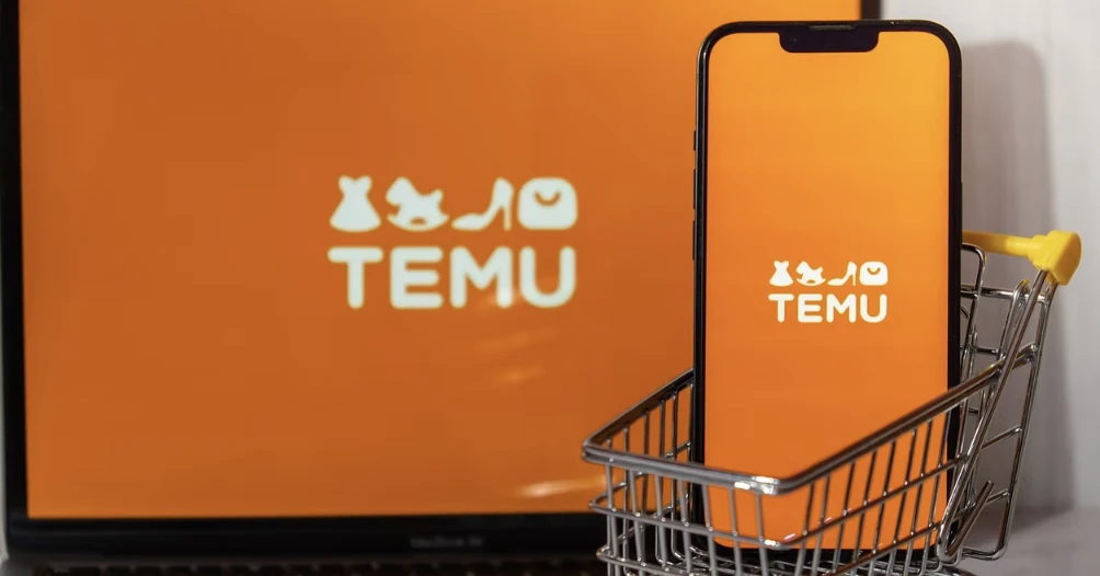 A phone showing the Temu logo, sitting in a small shopping cart in front of a laptop also showing the Temu logo